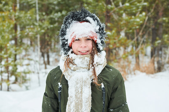 Portrait of Caucasian teenager boy smiling into camera with face and winter hat covered in snow standing outdoors in snowy forest during cold weather