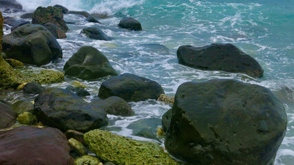 Waves crash against dark coastal rocks under turquoise seawater. Wet stones glisten in the sunlight, showing the raw beauty of a tropical shoreline.