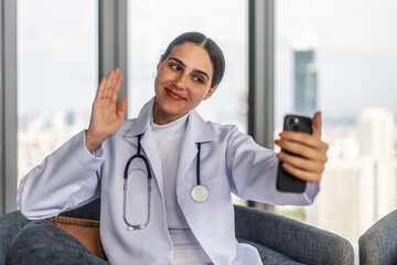 Young woman doctor is enjoying a friendly video call on her mobile phone with a patient or friend while sitting comfortably on a sofa. Female cheerful demeanor adds warmth to the cozy living room