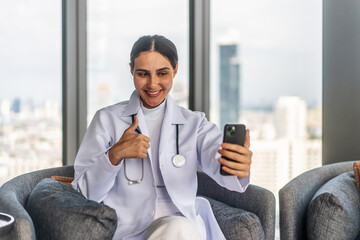 Young woman doctor is enjoying a friendly video call on her mobile phone with a patient or friend while sitting comfortably on a sofa. Female cheerful demeanor adds warmth to the cozy living room