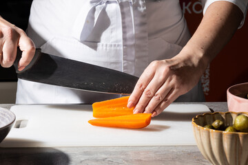 Chef in white uniform skillfully slices a carrot on a white cutting board with a large chef’s knife. Close-up of hands at work, with bowls of ingredients visible in the background. Focus on precision 