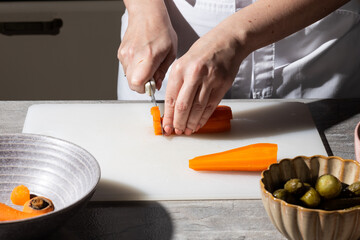 Close-up of hands slicing a carrot on a white cutting board. Ceramic bowls with sliced vegetables and pickles nearby. Clean kitchen setting, focus on precise food preparation.