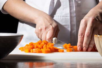 Close-up of hands dicing orange carrots on a white cutting board with a sharp knife. A bowl is ready to receive the chopped vegetables. Clean, professional kitchen scene, focus on food preparation.