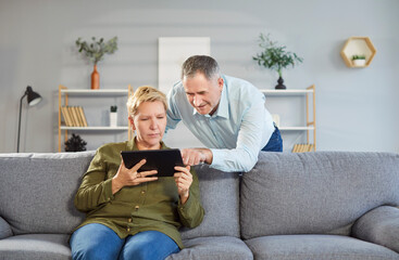 Portrait of elderly couple using digital tablet online entertainment or distant video call at home. Smiling senior man pointing at screen, showing something to his wife who sitting beside him on sofa.