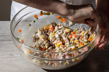 Close-up of hands mixing a classic Olivier salad in a large glass bowl. The salad features diced carrots, peas, potatoes, meat, and mayonnaise. Vibrant colors and textures highlight the traditional