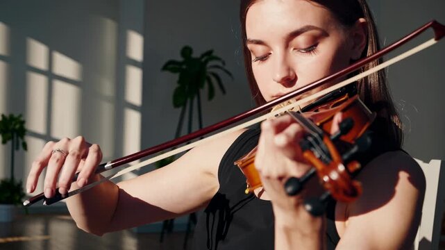 A serene video still of a woman playing the violin in a sunlit room. Captured from a side angle, emphasizing her focus and the warm ambiance.