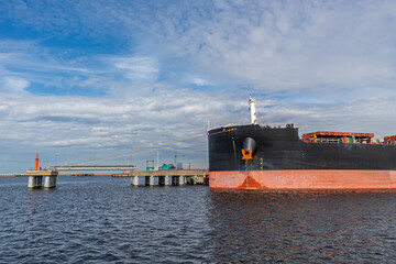 Large cargo ship docked at an industrial port terminal. Mooring cables are attached to the ship. Industrial port equipment and pipelines are visible in the background