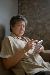 A Thoughtful elderly asian Woman Engaged Deeply with Her Smartphone While Relaxing in a Cozy Setting