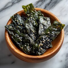 Top-down photo of crispy, seasoned seaweed snacks in a light-colored wooden bowl, which is set on a sophisticated white and grey marble countertop.