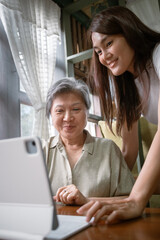Asian Thai woman patiently helps an elderly lady use a tablet, showcasing the beauty of connection and learning at cozy house.
