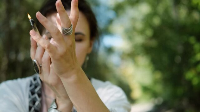 A close-up of a beautiful brunette holding a smoking palo santo stick during a magical ritual against a green forest backdrop. The woman, dressed in boho style, meditates and dances in the forest.