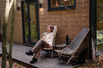 Young woman drinking tea on the veranda of a country house in the evening. Woman resting on the terrace of a house with a view of the forest. Vacation outside the city.
