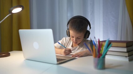 Young boy wearing headphones and using laptop. Child learning in distance. A kid is training his homework on a laptop. A young boy with headphones on, using lifestyle a laptop. - Powered by Adobe