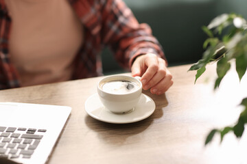 Coffee break. Woman with cup of aromatic drink and laptop at wooden desk indoors, closeup