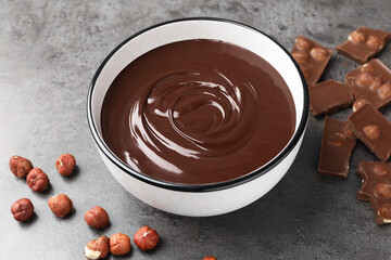 Melted chocolate in bowl, pieces and nuts on grey table, closeup