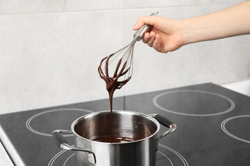 Woman holding whisk with melted chocolate above pot, closeup