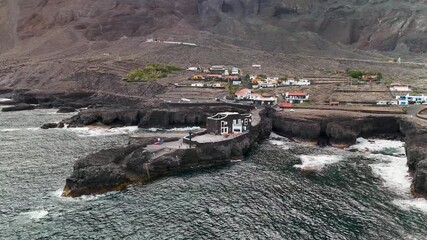 Circling aerial drone view of Hotel Punta Grande, recognized as the world's smallest hotel, perched dramatically on a volcanic pier in El Hierro, Canary Islands, Spain.
