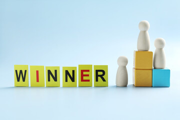 Cards with word Winner, human figures, wooden one on top of stacked cubes against light blue...