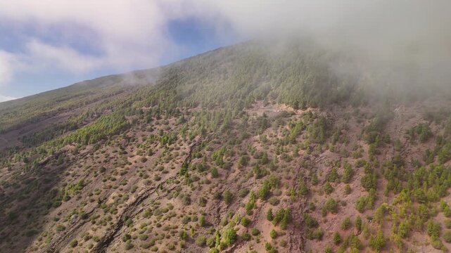 Rising aerial drone view over the volcanic slopes and ravines of La Restinga, capturing the dramatic layer of swirling clouds floating just above the Atlantic coast of El Hierro, Spain.