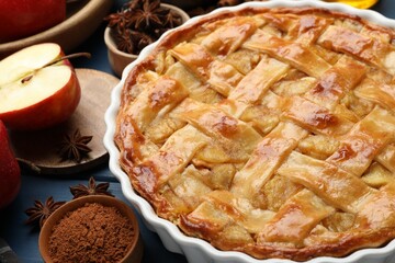 Delicious homemade apple pie in baking dish and ingredients on blue wooden table, closeup