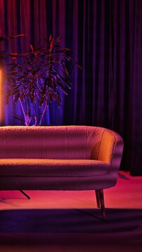 In a studio, a modern couch sits against dark curtains, illuminated by vibrant colored lights. The atmosphere is artistic and inviting, perfect for a photoshoot or video production.