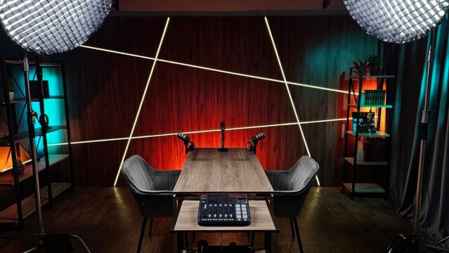 Two stylish chairs and a wooden table are set up in a modern studio, illuminated by dramatic lighting. Audio equipment is ready for podcasting sessions, creating a creative atmosphere.