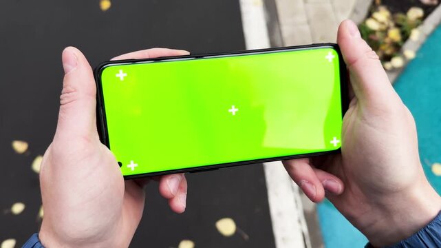 Handheld view of person walking and holding smartphone with green screen outdoors