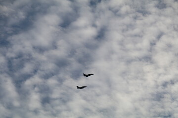 two small silhouettes of fighter jets fly in a blue sky with cumulus clouds