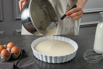 Woman pouring liquid dough from bowl into baking dish at grey table in kitchen, closeup