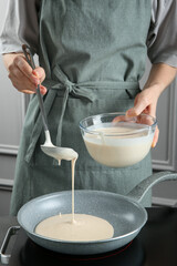 Woman pouring liquid dough from bowl into frying pan at grey table in kitchen, closeup