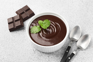 Melted chocolate with mint in bowl, pieces and spoons on light grey table, closeup