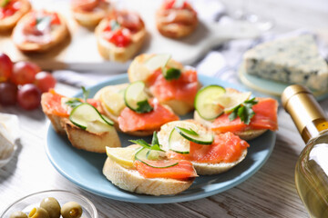 Tasty bruschettas with salmon, cucumber, lemon, olives and bottle of wine on light wooden table, closeup
