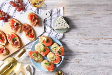 Tasty bruschettas with salmon, cucumber, lemon, prosciutto and wine on white wooden table, flat lay. Space for text