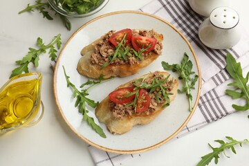 Delicious bruschettas with tuna, tomatoes and arugula served on white marble table, flat lay