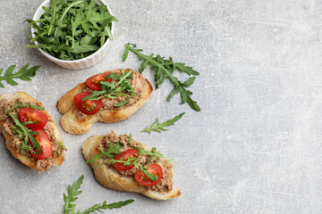Delicious bruschettas with tuna, tomatoes and arugula on grey table, flat lay. Space for text