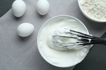 Whipped whites in bowl, whisk, flour and eggs on grey wooden table, flat lay