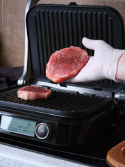 A man cooks marbled beef steaks on an electric grill against a backdrop of a home kitchen.