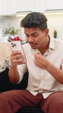 Indian man at home checks messenger long small text on smartphone puts on glasses and reads clearly. Arabian guy on sofa improves focus smiles and continues chat and scrolls through message thread