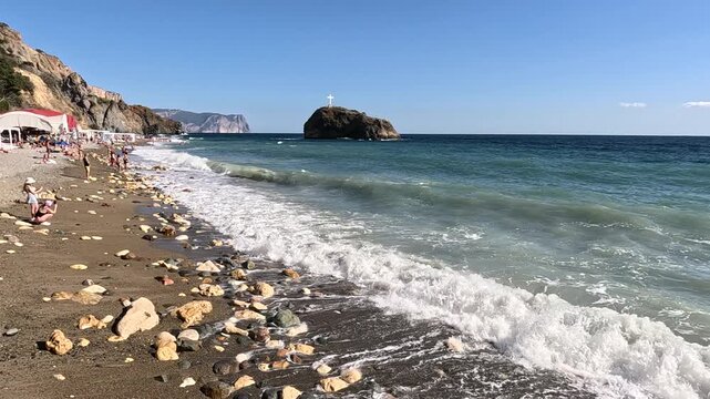 Beach waves sea, beautiful sunny day with gentle ocean waves crashing on a pebble shore, featuring a distant island and people.