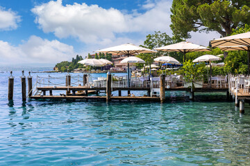 Relaxing by the sparkling waters of Sirmione at Lake Garda, meals on a wooden dock at Sirmione, surrounded by the blue waters of Lake Garda. Bright umbrellas provide shade on this sunny day.