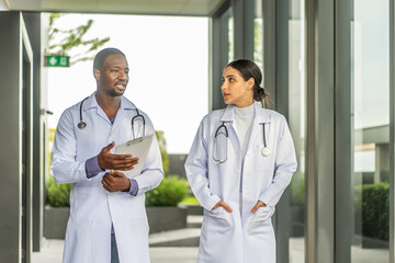 Two doctors man and woman walking and share a friendly conversation on the rooftop of a hospital during their lunch break. They discuss work and patient care while enjoying a sunny day in the city