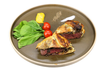 A delicious Beef Wellington served on a plate with fresh greens, mustard, and tomatoes. The wellington's crust is golden brown and flaky, revealing a tender, juicy interior
