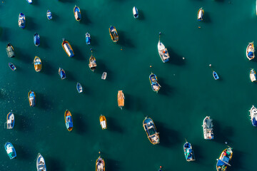 Aerial panorama shows dozens of Maltese luzzu boats anchored in Marsaxlokk, Malta. Bright hulls form oval patterns with shadows in clear daylight over teal water.