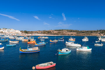 Mid day aerial view of Marsaxlokk Bay, Malta, with dozens of bright luzzu boats on calm blue water, whitewashed buildings and rocky shoreline framing the working harbor. © Aerial Film Studio