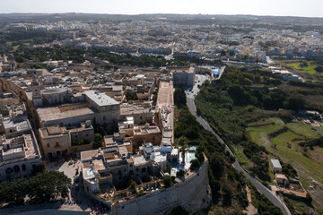 Fototapeta premium Aerial view shows Mdina, Malta, with honey colored walls, narrow streets, bastions, and main gate, midday light, visitors by the walls, Rabat and fields beyond