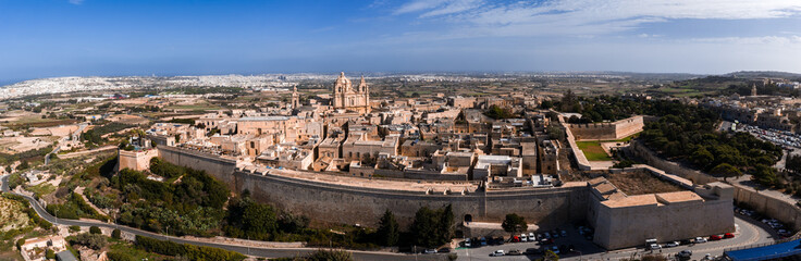 Fototapeta premium Aerial panorama shows Mdina, Malta, with limestone ramparts, St. Pauls Cathedral dome and bell towers, honey colored streets, clear daylight, and distant Mediterranean.