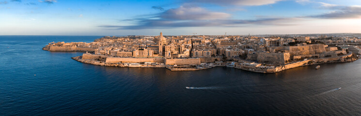 Obraz premium Aerial view of Valletta, Malta at sunset shows limestone bastions, Fort St. Elmo, the basilica dome, and cathedral spire as boats cross Marsamxett Harbour.