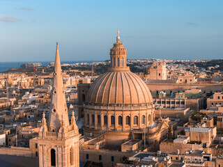 Obraz premium Aerial view of Valletta, Malta at sunset, the basilica dome and St. Pauls Pro Cathedral spire rise over honey colored limestone blocks, narrow streets, and the Mediterranean Sea.