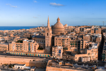 Fototapeta premium Aerial view of Valletta, Malta at sunset, basilica dome and cathedral spire rise above limestone streets and bastions, golden hour light warms sandstone beside the sea.