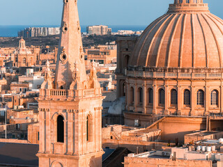 Aerial view shows Valletta, Malta, at golden hour, the basilica dome and Baroque bell tower in warm limestone above honey colored blocks, with modern apartments by the sea.
